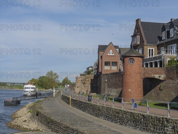 Historic brick buildings along a riverside footpath in sunny weather with a slightly cloudy sky,