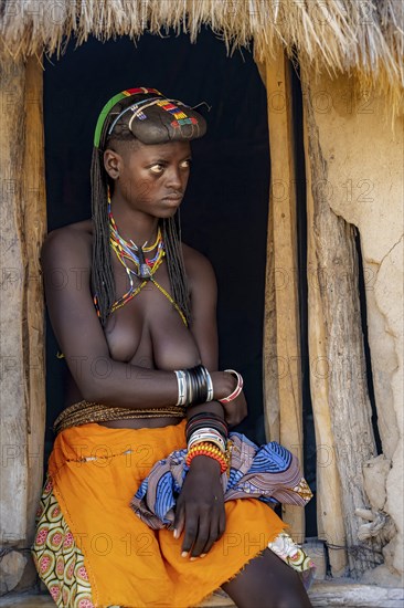 Hakaona woman with traditional kapapo hairstyle, sitting in the entrance of her mud hut, in the