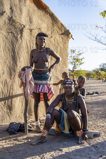Two Hakaona woman with traditional kapapo hairstyle, with children, in front of their mud hut, in