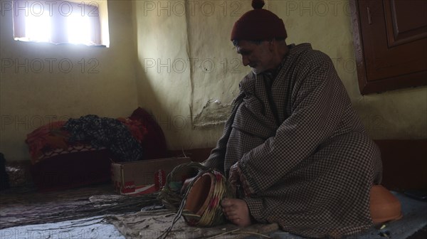 Elderly man in traditional clothing sitting in a dimly lit room with a basket and a contemplative
