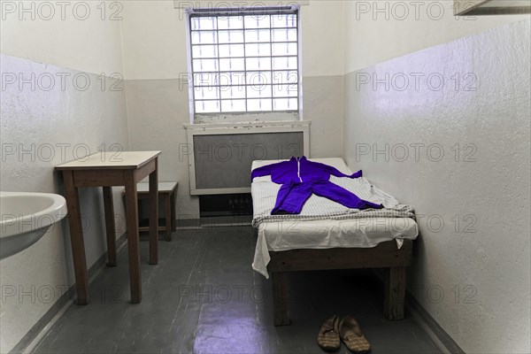 Simply furnished prison cell with bed and clothes. Small windows and cold lighting,