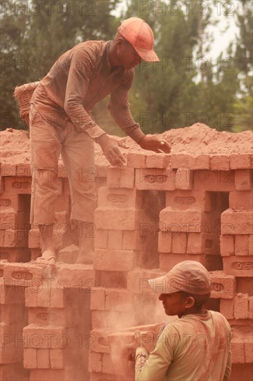 Two workers stacking red bricks in a dusty outdoor setting, both wearing hats and working together,