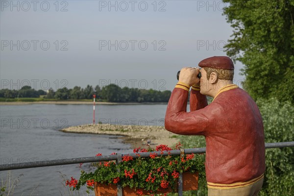 Man statue with binoculars on the riverside, flowers in the foreground and trees in the background,