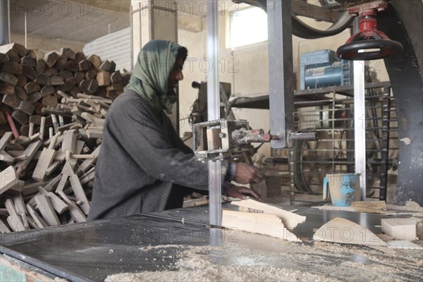 Man working in a wood workshop with machinery and logs around, wearing traditional clothing,
