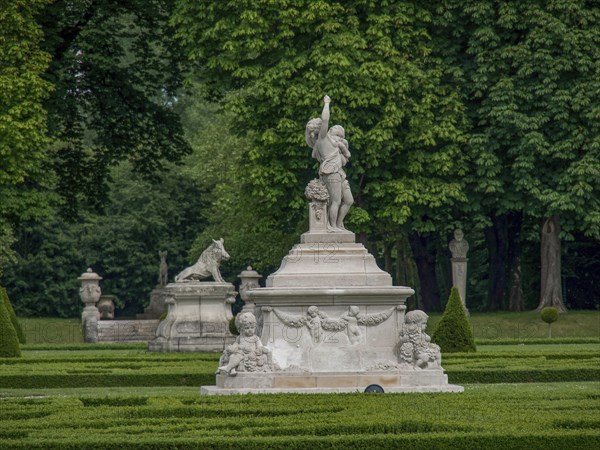Elegant stone figure in a lush, well-kept garden surrounded by green trees and hedges, old red