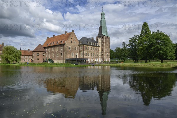 Large castle with brick walls and towers by a pond with reflections, surrounded by trees under a
