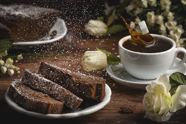 A cup of coffee, cake with icing sugar and splashing coffee drops, flower arrangement on a wooden