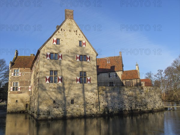 A historic moated castle with several windows and red shutters, on the banks of a pond,