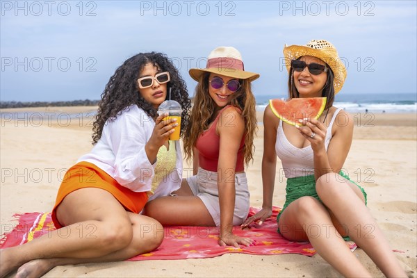 Three women sitting on a beach, one of them holding a watermelon slice. Scene is relaxed and