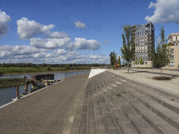 Riverbank with modern buildings in the background and a promenade, under a sky with small clouds,