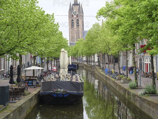 A calm river with boats and green trees on both sides, with a church tower in the background,