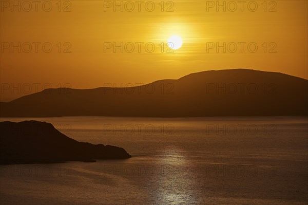 The sun sets over a calm sea and rolling hills, bathing the scene in warm orange, Makri Island,