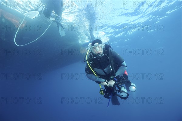A diver with a camera swims underwater next to a boat on the surface. Dive site Marine reserve Cap