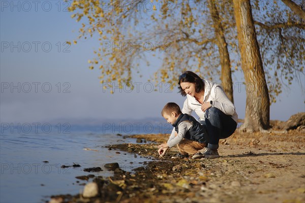 A mother and her child explore the shore of a calm lake during autumn surrounded by trees with