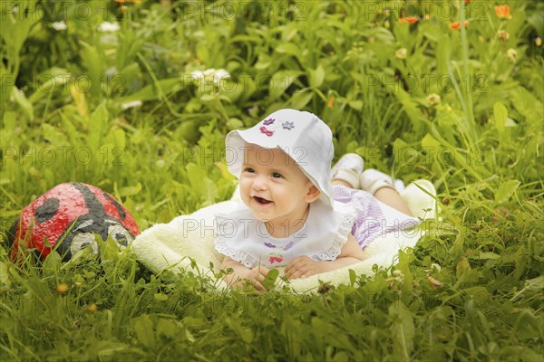 A smiling baby laying on the grass with a white hat, surrounded by flowers and greenery in the