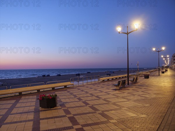 An empty promenade at dusk, illuminated by lanterns, the sea in the background, a peaceful and