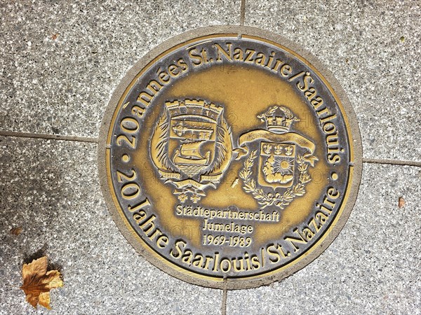 Bronze plaque embedded in a pavement. The plaque is in German and reads '20 Jahre St. Nazaire
