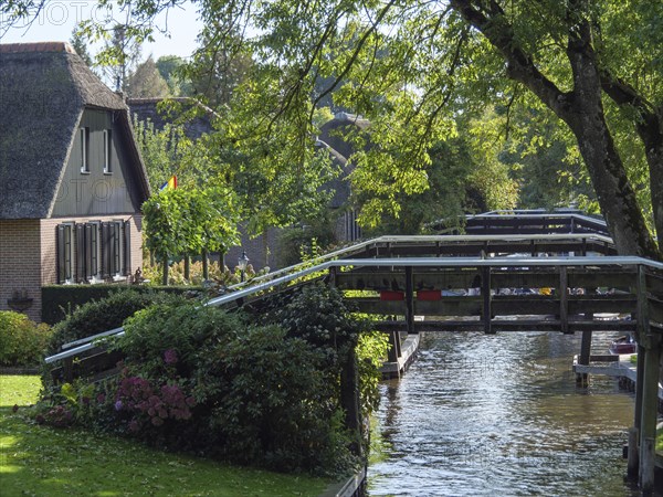 Wooden bridge over a canal, surrounded by trees and a house, with shadow effect through the leaves,