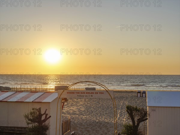 Sunset on the beach, quiet atmosphere, an archway with lettering, sea in the background, trees on