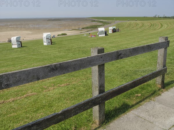 Meadow with beach chairs and a wooden fence along a path in front of a vast sandy beach and the sea