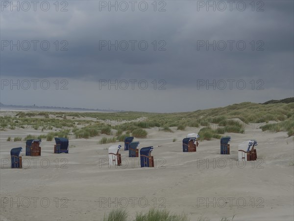 Scattered beach chairs on a sandy beach between grass and dunes under threatening clouds, Juist,
