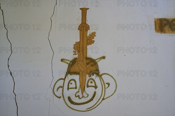 Graffiti of a face with a sword can be seen on a cracked white wall of an abandoned building, Lost