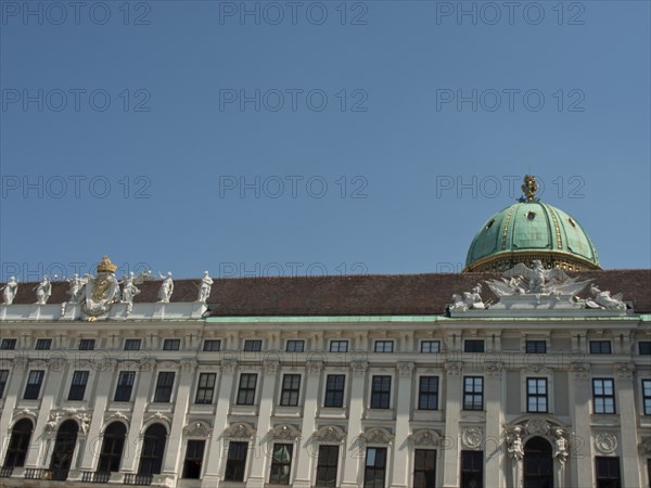 White baroque building with green domed roof and statues on the long façade under a blue sky,