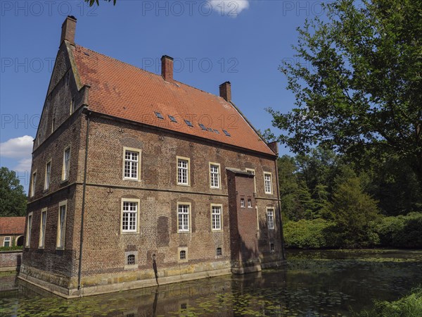 Old brick house with red roof on the edge of a pond, surrounded by green trees in summer, ochtrup,