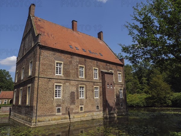Large brick building by a pond with calm water, surrounded by trees and sunny weather, ochtrup,