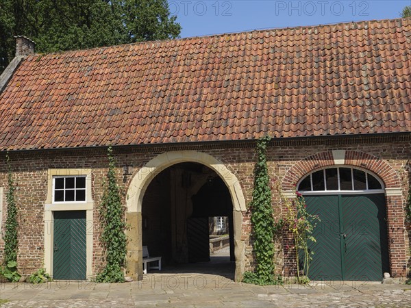A brick house with a round arch, red tiles on the roof, green doors and surrounded by climbing