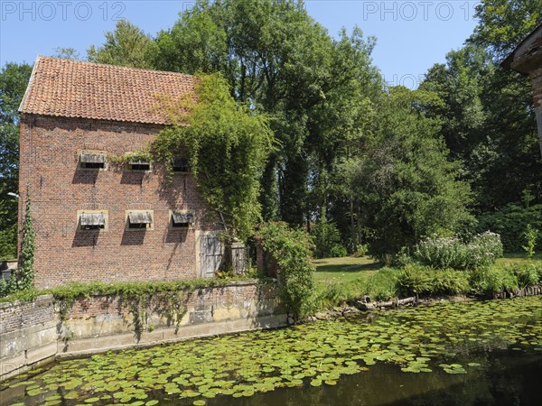 Red brick building with wooden shutters and ivy growth by the pond, surrounded by trees and sunny