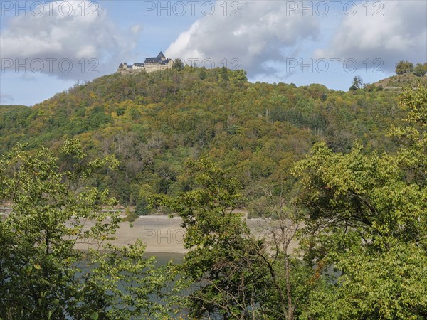 A majestic castle complex stands on a wooded hill that slopes down to a wide, river-like body of