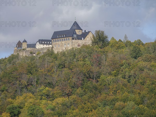 A medieval stone castle perched on a wooded hill surrounded by lush vegetation under a cloudy sky,