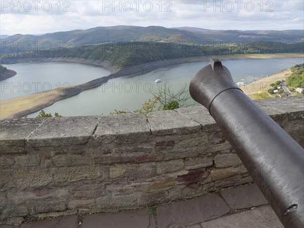 View from a stone wall with a cannon onto a lake surrounded by wooded hills under a cloudy sky,