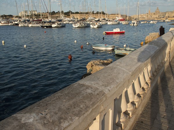 Stone balustrade along the harbour shore with many boats in the water and city in the background,