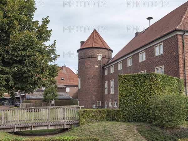 Brick building with tower and roof, surrounded by a hedge and trees, with a small bridge in the