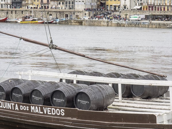 A boat loads wine barrels on the bank of a river while passers-by walk by in the background, Porto,