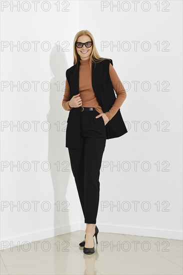 Minimalistic studio portrait of happy blonde woman in black vest, brown turtleneck and trousers