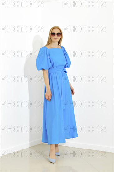 Beautiful woman with smooth blonde hair posing in bright room in long blue dress with short sleeves