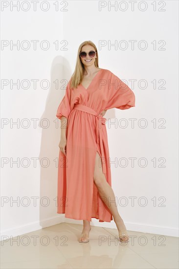 A fashion model showcases a floor-length coral dress with a v-neckline in a bright room