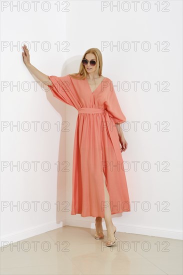 A stylish blonde woman in a coral maxi dress with a v-neckline leaning against white wall