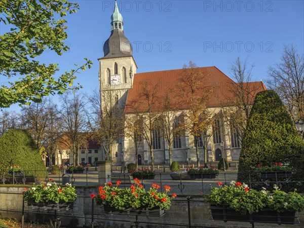 A historic church with a striking church tower, surrounded by blossoming flowers and autumnal trees
