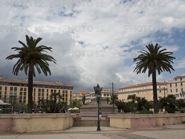 A large square with a statue in the centre, flanked by palm trees and surrounded by old buildings
