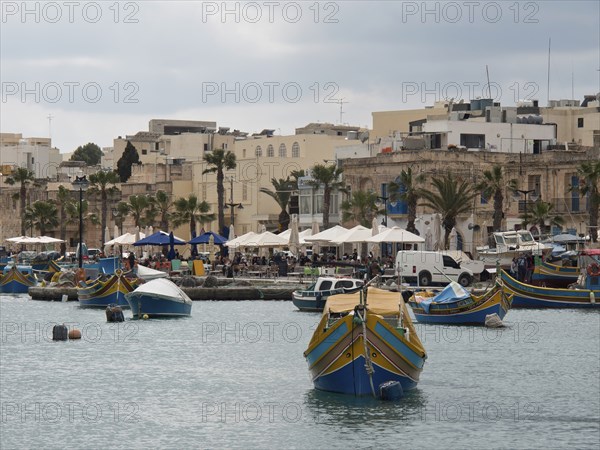 A row of fishing boats in front of a small town with palm trees, under a cloudy sky, marsaxlokk,
