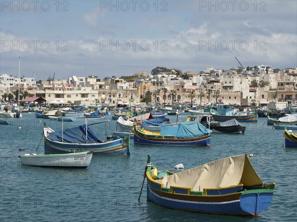 A harbour full of fishing boats with buildings in the background under a cloudy sky, marsaxlokk,