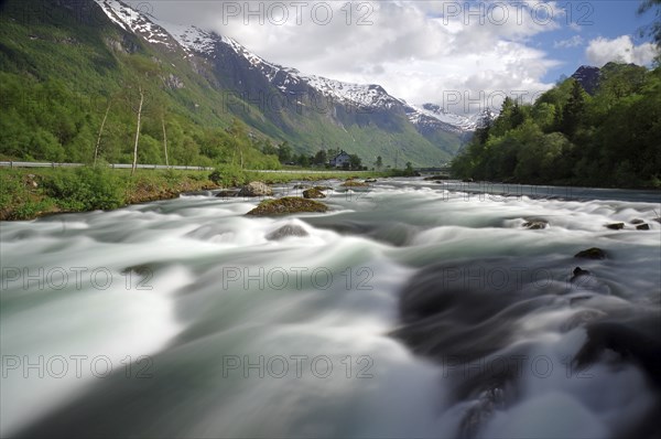 A wide river in a mountainous landscape with snow-capped peaks and cloudy skies, Olden, Nordfjord,