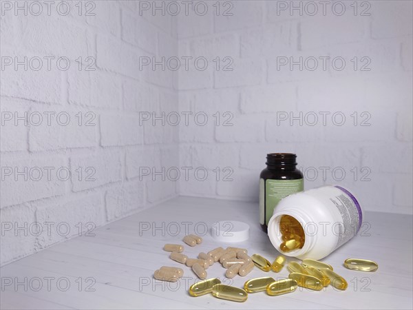 Two bottles of vitamins with spilled pills on a white surface against a white brick wall