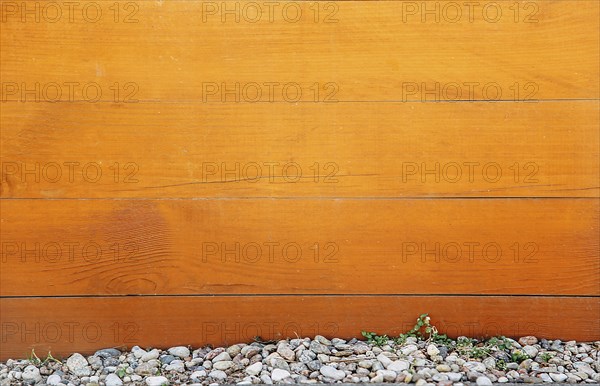 Wooden fence with orange-brown planks, pebbles and rocks at the bottom, a small green plant growing