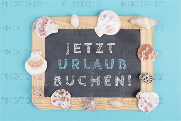Book vacation now is standing in german language on the chalkboard, seashells bulit frame, holiday