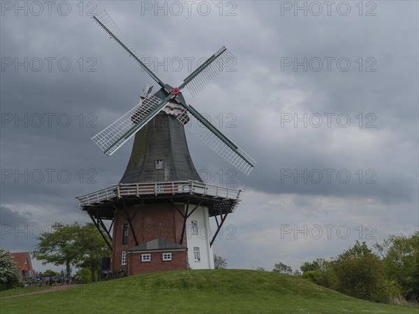 Old windmill on a green hill in front of a cloudy sky and a brick white building, greetsiel, east